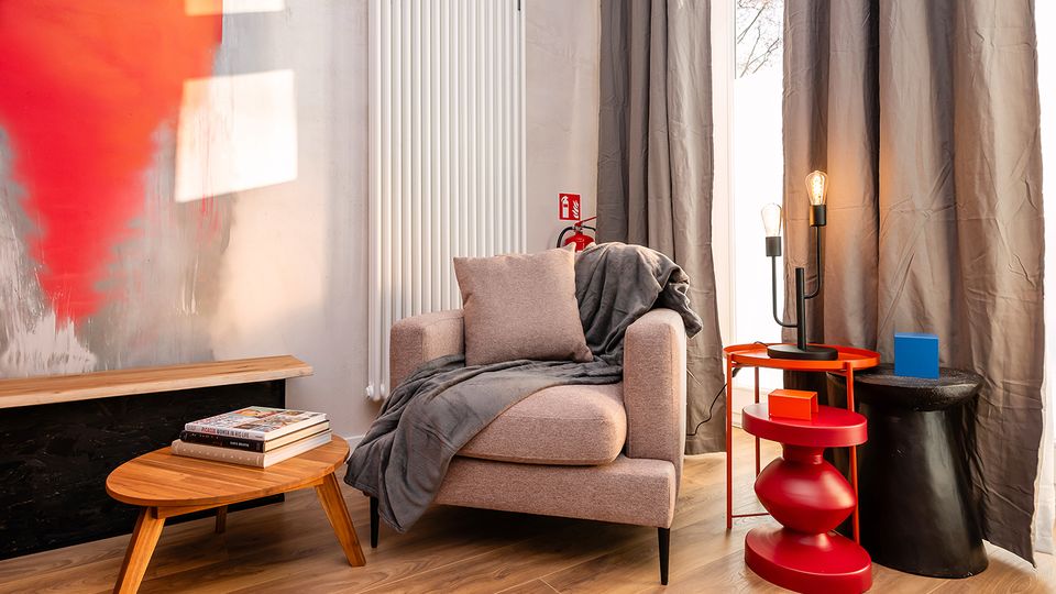 Gemütliche Wohnecke mit beigem Sessel und grauer Decke neben einem großen Fenster mit langen grauen Vorhängen. Davor stehen ein niedriger Holztisch mit Büchern sowie farbige Beistelltische mit Lampen; an der Wand befinden sich ein weißer Heizkörper und ein abstrakter roter Wandbereich.