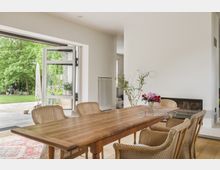 Helles Esszimmer mit einem großen Holztisch und geflochtenen Stühlen, geöffneten Glastüren zum Garten, weiße Wände und einem Kamin an der rechten Seite. Durch die Tür sind eine Terrasse und eine grüne Rasenfläche mit Bäumen sichtbar.