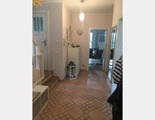 Ein heller Flur in einem Wohnhaus mit Fliesenboden, einer weißen Eingangstür, einem Treppenaufgang auf der linken Seite und einer schmalen Kommode mit einer Laterne darauf. Im Hintergrund ist eine Türöffnung, die in ein Esszimmer mit Esstisch und Stühlen führt.