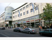 Straßenansicht eines Einkaufszentrums, der "Steglitzer Arkaden" in Berlin, mit einer modernen Gebäudefassade aus hellem Stein, großen Fensterfronten und einem runden Glasgebäude auf der linken Seite. Im Erdgeschoss befinden sich verschiedene Geschäfte und ein Eingang, während davor geparkte Autos sowie Fußgänger auf dem Bürgersteig zu sehen sind.