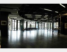 Großer, moderner Innenbereich mit glatten Steinfliesen, Glaswänden und Deckenleuchten. Im Hintergrund befinden sich bodentiefe Fenster und Glastüren, die den Blick nach draußen ermöglichen, sowie Sitzbänke und dekorative Wandbilder.