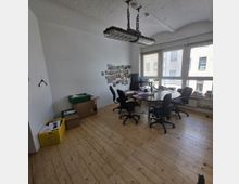 Ein einfaches Büro mit Holzboden, weiß gestrichenen Wänden und einer großen Fensterfront, die viel Tageslicht hereinlässt. Im Raum stehen ein runder Besprechungstisch mit mehreren Bürostühlen, ein Computerarbeitsplatz sowie Regale und Kartons; an der Wand sind Fotos und Notizen angebracht.