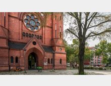 Backstein-Kirche mit großen gotischen Bögen und einem kreisförmigen Rosettenfenster über dem Eingang, umgeben von Bäumen und einem offenen Platz mit Pflastersteinen; daneben ist ein mehrstöckiges Wohngebäude zu sehen.