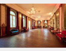 Großer, historisch anmutender Salon mit hohen Decken, Kronleuchtern und großen, bogenförmigen Fenstern, die von roten Vorhängen eingerahmt sind. Der Raum ist mit antiken Möbeln wie roten und blauen Sofas sowie einem Klavier im Hintergrund ausgestattet.