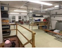 Ein Lagerraum mit Regalsystemen, die Kisten, Materialien und Gegenstände wie Leitern und Rohre enthalten. Im Hintergrund sind eine Glastrennwand sowie ein Bereich mit Sitzmöbeln und Tischen zu sehen, während die Decke mit sichtbaren Kabeln und Hängelampen ausgestattet ist.