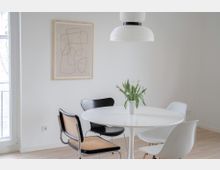 Heller Essbereich mit einem runden weißen Tisch, umgeben von vier verschieden gestalteten Stühlen, dekoriert mit einer Vase voller weißer Tulpen. Im Hintergrund ein abstraktes Bild an einer weißen Wand und eine moderne, weiße Pendelleuchte.