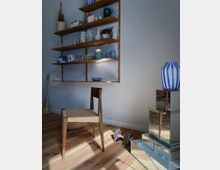 Holzstuhl mit geflochtener Sitzfläche in einem hellen Raum mit Holzboden, daneben eine Wand mit offenen Holzregalen, die Keramik, Geschirr und Dekorationsgegenstände beherbergen. Im Vordergrund steht ein gestapeltes Regal aus spiegelnden Würfeln mit einer blau-weiß gestreiften Vase darauf, während Tageslicht durch den Raum fällt.