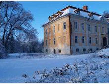 Historisches Herrenhaus mit gelber Fassade und weißen Fensterrahmen, umgeben von einer schneebedeckten Landschaft mit kahlen Bäumen im Hintergrund. Klarer blauer Himmel ergänzt die winterliche Stimmung.