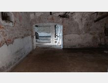 Ein leerer Kellerraum mit rohen Backsteinwänden, teils verputzt, und einem gewölbten Deckenbogen. Im Hintergrund ist ein weiterer Raum mit freiliegendem Mauerwerk und einigen herumliegenden Gegenständen sichtbar.