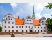 Historisches Schlossgebäude mit weißer Fassade, roten Ziegeldächern und Renaissancegiebeln, ergänzt durch einen grünen Kirchturm im Hintergrund; gepflasterter Vorplatz mit Blumenschmuck und umliegendem Baumbewuchs.