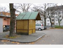 Ein kleiner hölzerner Unterstand mit einem grünen Satteldach steht auf einem gepflasterten Parkplatz. Im Hintergrund sind mehrstöckige Wohnhäuser, kahle Bäume und geparkte Autos sichtbar.