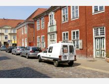 Straßenszene mit Kopfsteinpflaster, gesäumt von roten Backsteinhäusern mit weißen Fensterrahmen und grünen Holztüren; parkende Autos, darunter ein weißer Lieferwagen, stehen entlang des Gehwegs. Im Hintergrund ein helles Gebäude mit Satteldach.