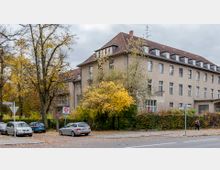 Mehrstöckiges Wohn- oder Bürogebäude mit grauer Fassade und einem roten Satteldach, umgeben von Bäumen mit herbstlichem Laub. Im Vordergrund sind geparkte Autos, eine Straße mit Tempo-30-Schild und Fußgänger zu sehen.