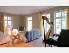 Wohnzimmer mit Holzboden und großen Fenstern, die mit gelben Vorhängen dekoriert sind. Der Raum ist eingerichtet mit einem gestreiften Sofa, einer Stehlampe, einem kleinen Fernseher, einer Harfe und einem Klavier.