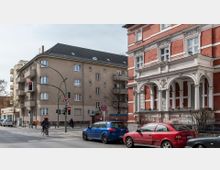 Straßenkreuzung in einer Stadt mit einem historischen Backsteingebäude rechts, das weiße Verzierungselemente und Arkaden aufweist, sowie einem schlichten Wohngebäude links. Mehrere geparkte Autos, Fahrradfahrer und eine Ampel sind im Vordergrund sichtbar.