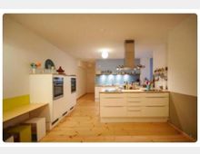 Moderne Küche mit hellen Holzböden, einer zentralen Kücheninsel mit integriertem Herd und Abzugshaube sowie weißen Schränken. Im Hintergrund befinden sich ein doppelt eingebauter Backofen und Regale vor einer hellblauen Wand.