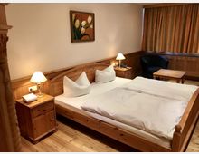 Ein gemütlich eingerichtetes Hotelzimmer mit Holzmöbeln, darunter ein Doppelbett mit weißen Bettdecken, zwei Nachttische mit Lampen und ein kleiner Tisch mit einem Sessel. An der Wand hängt ein Gemälde mit Tulpen, und die Fenster sind mit Vorhängen versehen.