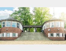 Backsteingebäude mit zwei runden, symmetrischen Türmen und großen Fenstern, verbunden durch einen überdachten Durchgang; davor eine Treppe und ein sandiger Boden. Im Hintergrund befinden sich hohe grüne Bäume.