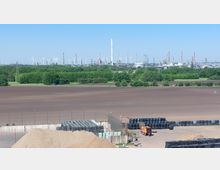 Weitläufige Landschaft mit einem brachliegenden Feld im Vordergrund, einer grünen Baumreihe in der Mitte und einer Industriekulisse im Hintergrund, die aus Windkraftanlagen, Schornsteinen und Tankbehältern besteht; im Vordergrund sind zudem gestapelte schwarze Ballen und eine orangefarbene Maschine sichtbar.