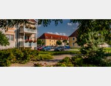 Wohnanlage mit gelb gestrichenen Mehrfamilienhäusern, roten Ziegeldächern und Balkonen mit Blumenkästen. Im Vordergrund befinden sich ein gepflegter Garten mit Sträuchern und Bäumen sowie ein Parkplatz mit mehreren Autos.