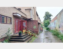 Ein Eingang zu einem Gebäude mit roten Türen und einer Backsteinmauer, flankiert von Pflanzen in Töpfen auf einer kleinen Treppe. Rechts verläuft ein Weg entlang einer bemalten Wand, im Hintergrund sind ein Zaun und Bäume zu sehen.