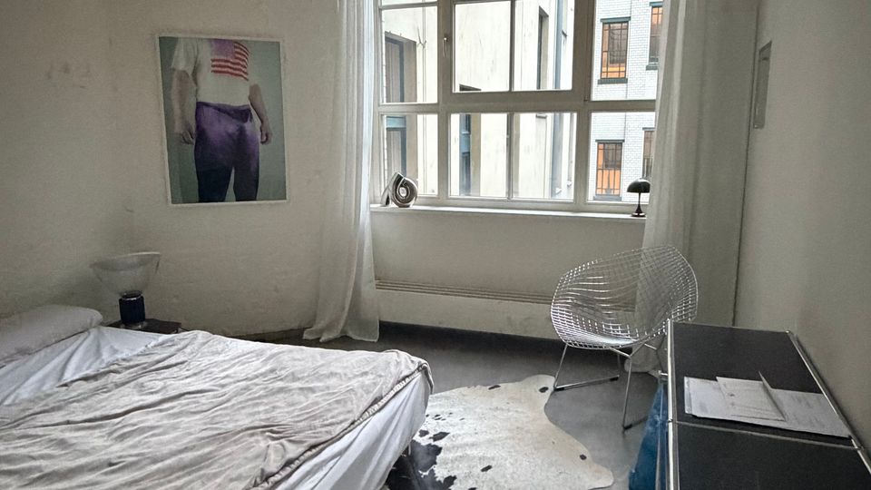 Schlichtes Schlafzimmer mit großem Sprossenfenster und weißen Vorhängen, Blick in einen Innenhof zwischen mehrstöckigen Gebäuden. Links ein niedriges Bett, auf dem Boden ein helles Kuhfell, rechts ein kleiner Schreibtisch mit transparentem Stuhl und Kommode.