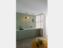 Minimalistisch gestaltete Küche mit grüner Wand und Schrankfronten, schwarzer Armatur und einer Arbeitsplatte. Drei Hängeleuchten mit Glaslampenschirmen hängen von der Decke, während ein goldener Tisch mit einer Glasdekorschale im Vordergrund steht.