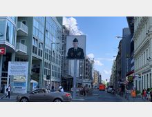 Straßenszene am Checkpoint Charlie in Berlin mit einem großen Porträt eines Soldaten auf einem Schild in der Mitte. Umgeben von modernen und historischen Gebäuden, sind Schilder eines Museums, Restaurants sowie ein Corona-Testzentrum sichtbar.