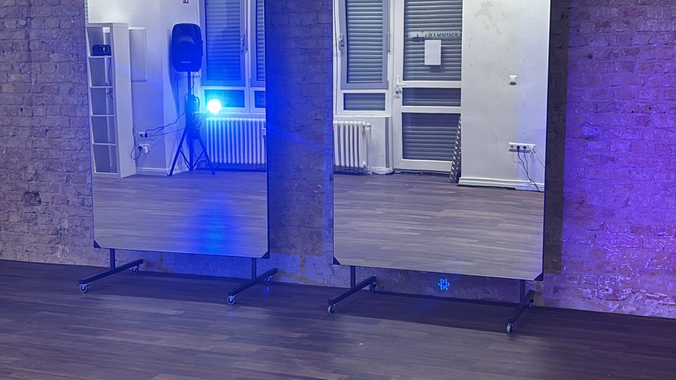 Dance or fitness studio with a wooden floor and an exposed brick wall, featuring two large freestanding mirrors on wheeled stands. Reflections show tall windows with blinds, a radiator, and studio equipment, with blue lighting cast across the room.
