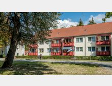 Mehrfamilienhaus mit cremefarbener Fassade und roten Balkonen, umgeben von Bäumen und einer Straße, auf der eine Person mit einem Fahrrad fährt. Der Vorgarten ist gepflegt, mit niedrigen Büschen und Blumen schmücken die Balkone.