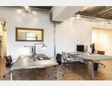 Moderner Büroraum mit weißen Wänden, freiliegender Verkabelung an der Decke und mehreren Schreibtischen mit Bürostühlen. Auf den Tischen befinden sich Monitore, Lampen und Tastaturen, während ein großes Holzfenster einen Blick in einen angrenzenden Raum bietet.