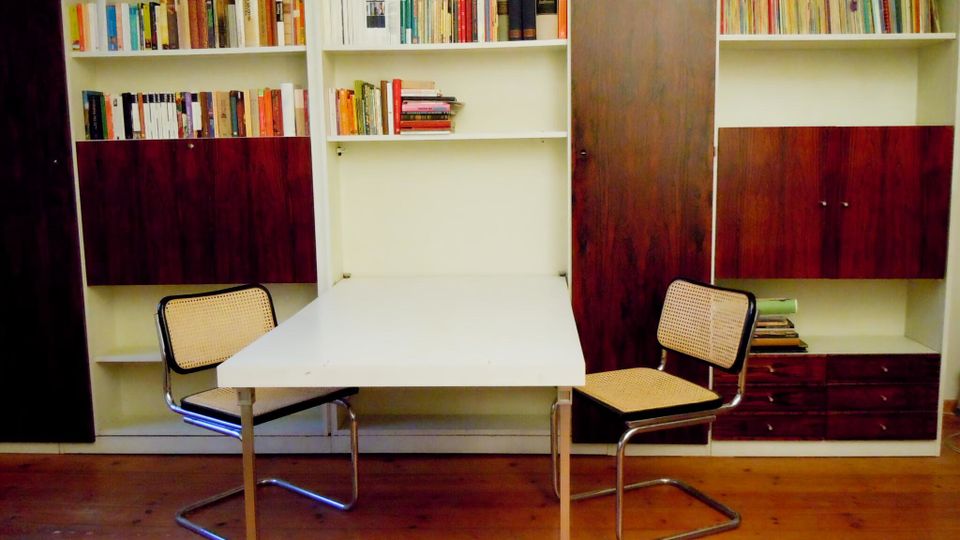 Arbeits- oder Leseecke mit weißem Tisch und zwei Freischwinger-Stühlen vor einer wandfüllenden Einbau-Bücherwand. Die Regale sind mit Büchern gefüllt und haben dunkelholzfarbene Schranktüren und Schubladen über einem Holzboden.