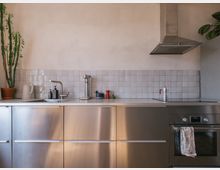 Moderne Küche mit Edelstahl-Unterschränken, einer weißen Arbeitsplatte und einem Fliesenspiegel aus kleinen, glänzenden, weißen Fliesen. Sichtbar sind ein Spülbecken, eine Dunstabzugshaube aus Edelstahl, ein Induktionskochfeld, ein Einbaubackofen sowie eine dekorative Pflanze in einem Terrakottatopf.