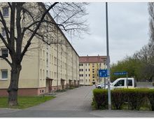 Mehrstöckige Wohngebäude in einem hellen Beigeton mit roten Dächern säumen eine gepflasterte Straße. Im Vordergrund steht ein Baum, rechts ist ein Straßenschild mit der Aufschrift „Berliner Str. 60 a–e“, daneben geparkte Fahrzeuge und grüne Büsche.