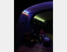 Ein dunkler Raum mit abgerundeten Wänden und einer Holzdecke, beleuchtet durch bunte LED-Streifen in Blau und Gelbgrün entlang der Deckenränder. Im Vordergrund befinden sich zwei Personen, die auf einem antik wirkenden, braunen Ledersofa sitzen, daneben ein kleiner Tisch mit Gegenständen.