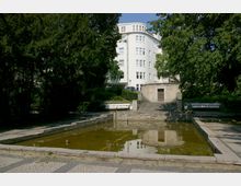Ein kleiner, rechteckiger Wasserbeckenbereich in einem parkähnlichen Umfeld, umgeben von Pflastersteinwegen, Bänken und üppigen grünen Bäumen im Hintergrund. Dahinter steht ein mehrstöckiges, weißes Gebäude mit abgerundeten Ecken und zahlreichen Fenstern.