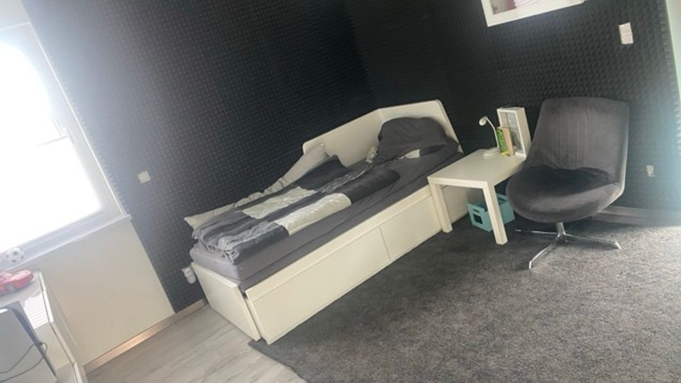 Schlafzimmer mit weißem Tagesbett und grauer Bettwäsche vor dunkel verkleideten Wänden, daneben ein kleiner weißer Beistelltisch und ein grauer Drehsessel. Links fällt Tageslicht durch ein Fenster ein; der Boden ist teils mit hellem Holz und teils mit einem großen grauen Teppich ausgelegt.