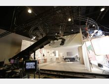 Filmstudio mit einer Wohnzimmerkulisse, ausgestattet mit Sofa, Sessel, Couchtisch und minimalistischer Dekorationsgestaltung. Im Vordergrund Kameratechnik und Beleuchtung, einschließlich eines Kamerakrans und Scheinwerfern, sowie technische Monitore.