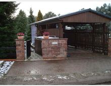 Holzüberdachter Carport mit angrenzender Einfahrt, flankiert von zwei gemauerten Säulen mit Blumenkübeln. Im Hintergrund sind Bäume sowie ein kleiner Gartenbereich sichtbar, teils schneebedeckt.