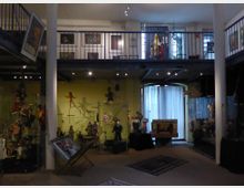 Ein Innenraum eines Museums mit Puppen- und Marionettenausstellungen. Die Wände sind mit Vitrinen, hängenden Figuren und Bildern dekoriert, während eine zweite Etage mit Geländer und Bildern sichtbar ist. Im Hintergrund befindet sich ein großes Fenster mit halbtransparenten Vorhängen.