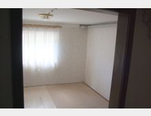 Leerer Raum mit weißen Tapeten, hellem Bodenbelag und einem Fenster mit durchscheinendem weißen Vorhang. An der Decke hängt eine einfache Lampe, und in der Ecke des Raumes sind Heizungsrohre sichtbar.