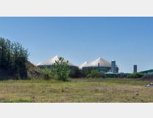 Biogasanlage mit mehreren großen, kuppelförmigen Silos im Hintergrund, umgeben von Bäumen und Vegetation auf einem offenen, grasbewachsenen Gelände. Der Himmel ist klar und blau.