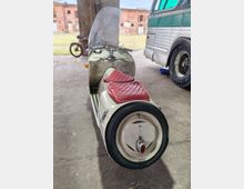 Überdachter Industriehof oder Lagerhalle mit Betonboden und Stützpfeiler, davor eine offene Rasenfläche und alte Backsteingebäude. Im Vordergrund steht ein Motorroller mit Beiwagen, daneben ist die Seite eines großen Busses sichtbar.