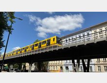 Gelber U-Bahn-Zug fährt über eine erhöhte Stahlbrücke vor dem Hintergrund eines blauen Himmels mit wenigen Wolken; darunter ältere Gebäude mit heller Fassade und geparkte Autos.