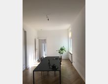 Ein heller Raum mit weiß gestrichenen Wänden, einer einfachen Hängelampe an der Decke und einem Holzboden in Fischgrätmuster. In der Mitte steht ein schwarzer Tisch mit Stühlen und darauf liegen verschiedene Gegenstände; ein Fenster und eine Zimmerpflanze befinden sich im Hintergrund.