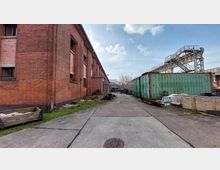 Industriegelände mit einem langen Ziegelgebäude auf der linken Seite und mehreren rostigen Containern auf der rechten Seite. Der Boden besteht aus Beton und im Hintergrund sind vereinzelte Bäume und weitere Gebäude sichtbar.