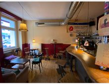 Gemütlicher Bar- und Caféinnenraum mit Holzfußboden, kleiner Sitzecke am Fenster, gemischten Tischen und Hockern sowie einer geschwungenen Theke. Sichtbare Lüftungsrohre an der Decke, Hängelampen und Regale mit Flaschen hinter der Bar prägen das Interieur.