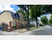 Wohnstraße mit gepflasterter Fahrbahn, gesäumt von Bäumen und Einfamilienhäusern in traditioneller Bauweise mit Gärten. Im Vordergrund ein gelb gestrichenes Haus mit schwarzem Walmdach, rotem Klinkerzaun und lila blühendem Strauch.