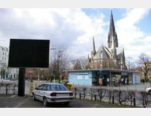 Ein städtischer Platz mit einer U-Bahn-Station ("U-Bahnhof Südstern") im Vordergrund und einer historischen Kirche mit spitzen Türmen im Hintergrund. Links steht ein unbeschriftetes, schwarzes Werbeschild, daneben ein silbernes Auto auf einem Parkplatz.