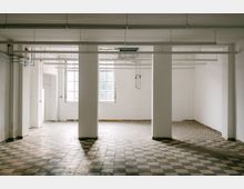 Leerer, heller Industrie- oder Lagerraum mit weiß gestrichenen Wänden, vier rechteckigen Stützpfeilern und einem schwarz-weiß karierten Fliesenboden. An der Decke verlaufen lange Leuchtstoffröhren und Leitungen; hinten sind große Fenster, die Tageslicht hereinlassen.